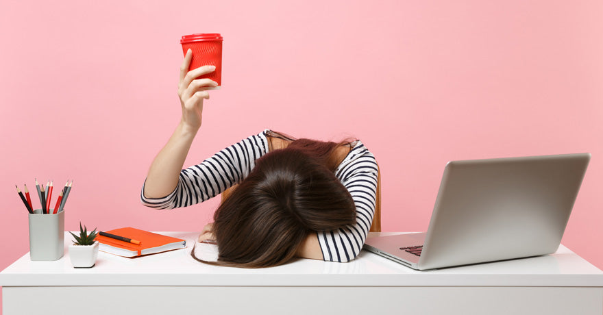 Woman with cup raised, head on table next to computer